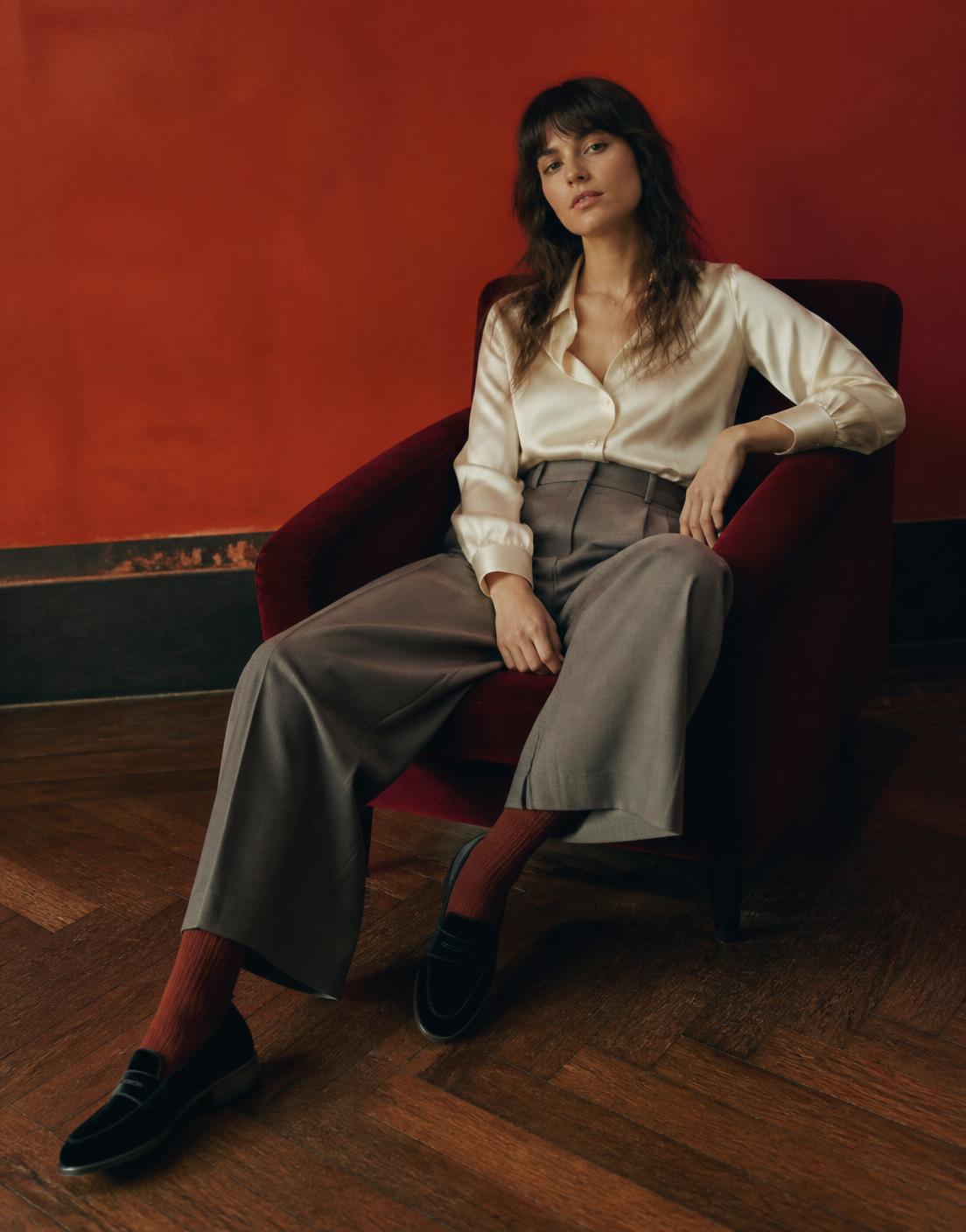 Model seated against a vermilion wall wearing black velvet penny loafers